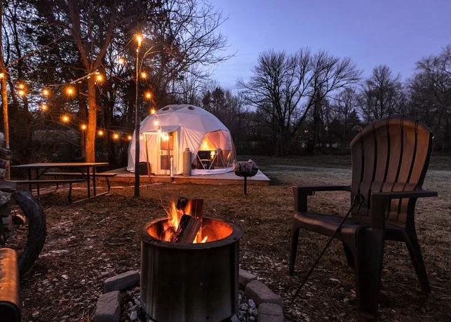 Dôme géodésique blanc éclairé le soir, entouré de guirlandes lumineuses, avec un feu de camp et des chaises en bois pour une ambiance chaleureuse en pleine nature.